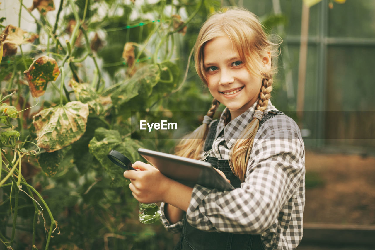 A beautiful little teenage girl with a tablet in her hands examines a sample of a plant through 