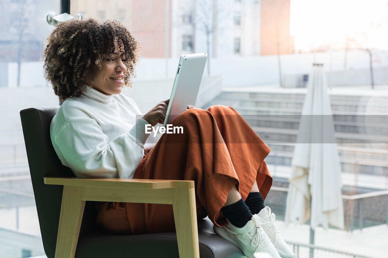 Side view of content african american female sitting in comfortable armchair and browsing tablet while entertaining during weekend at home