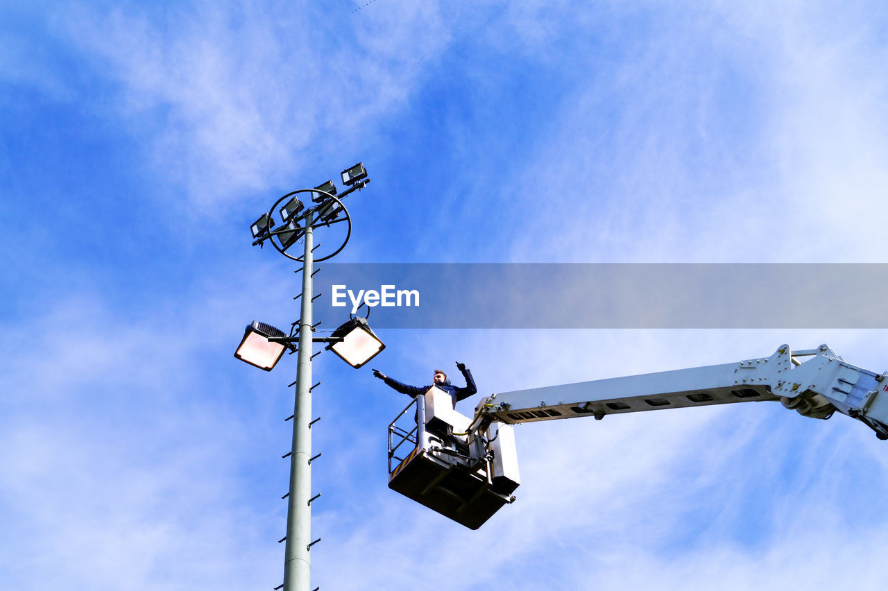 Low angle view of man standing on cherry picker by floodlight against sky