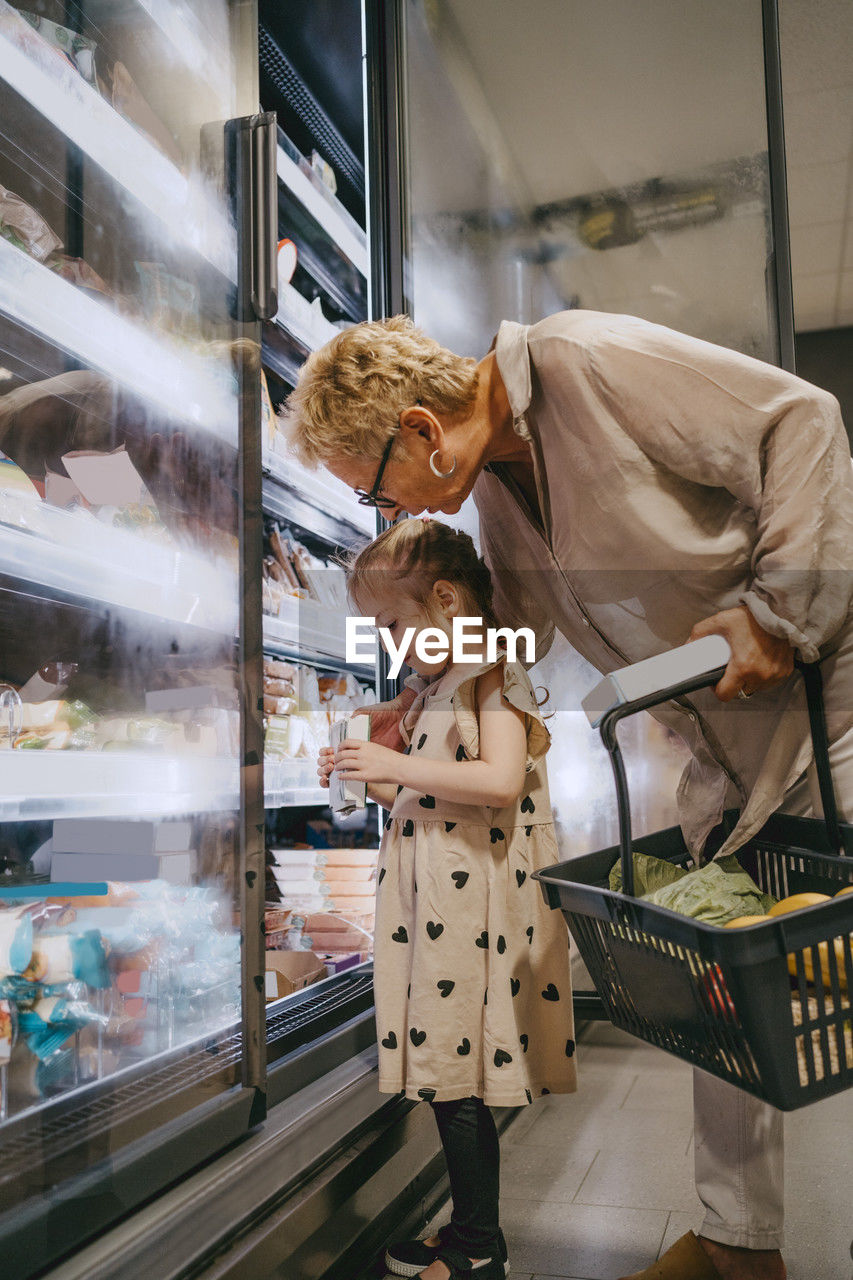 Senior woman with granddaughter choosing frozen food while doing shopping at grocery store