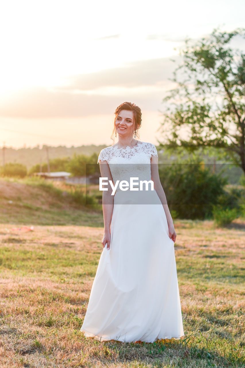 Woman wearing wedding dress while standing on field against sky during sunset