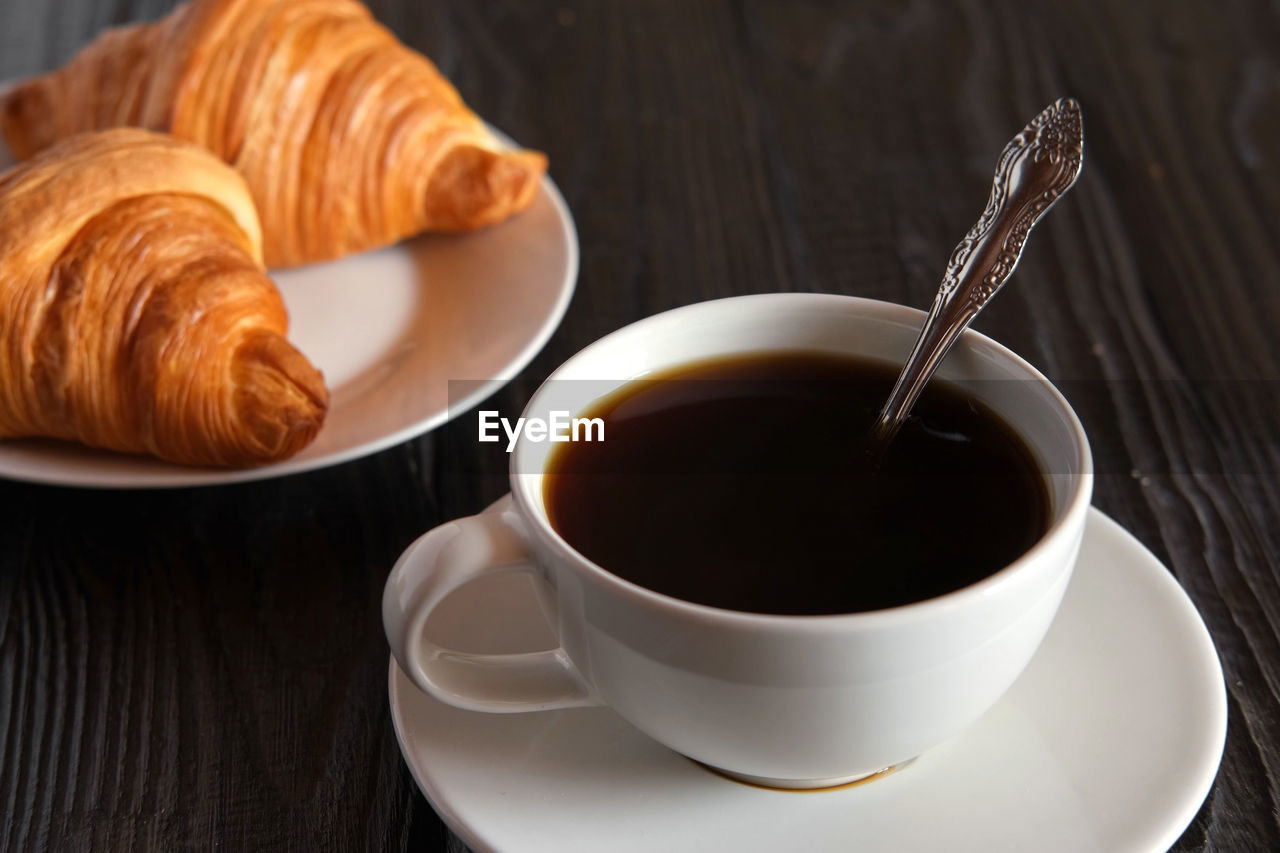 croissant, food and drink, cup, drink, mug, food, french food, coffee, coffee cup, refreshment, tableware, meal, baked, crockery, breakfast, hot drink, pastry, dessert, kitchen utensil, baked pastry item, fast food, sweet food, freshness, eating utensil, table, spoon, saucer, tea, no people, brown, close-up, indoors, cafe, wood, tea cup