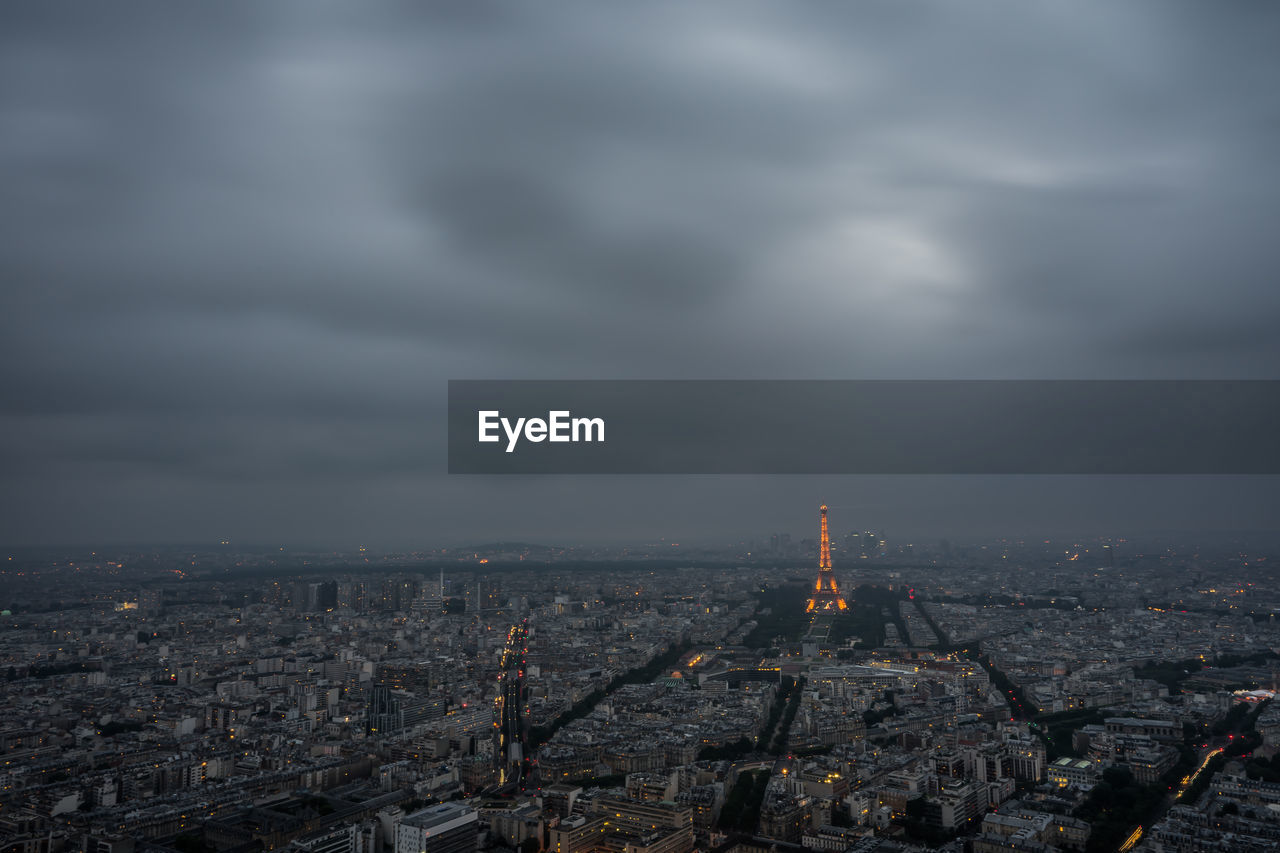 Distant view of eiffel tower amidst cityscape against cloudy sky at dusk