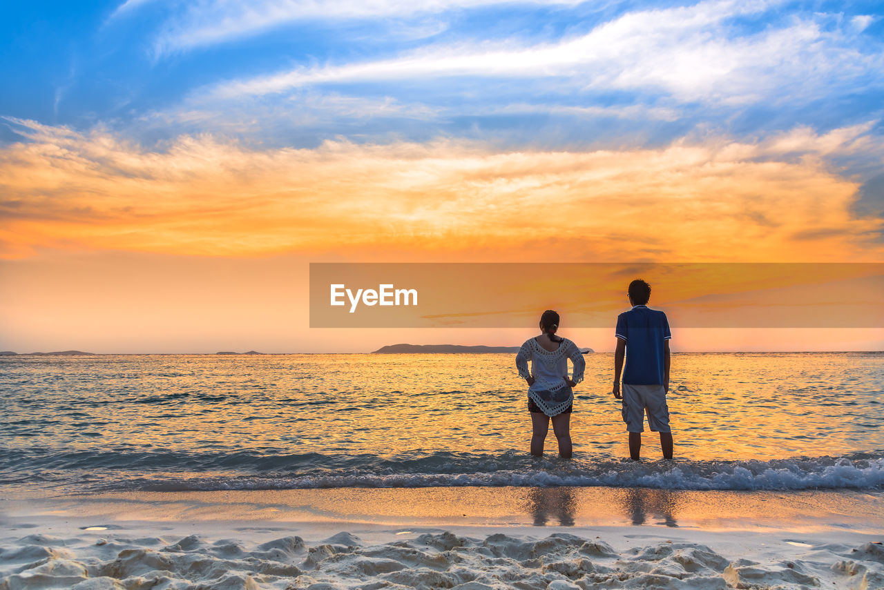 Rear view of couple standing on shore at beach against sky during sunset