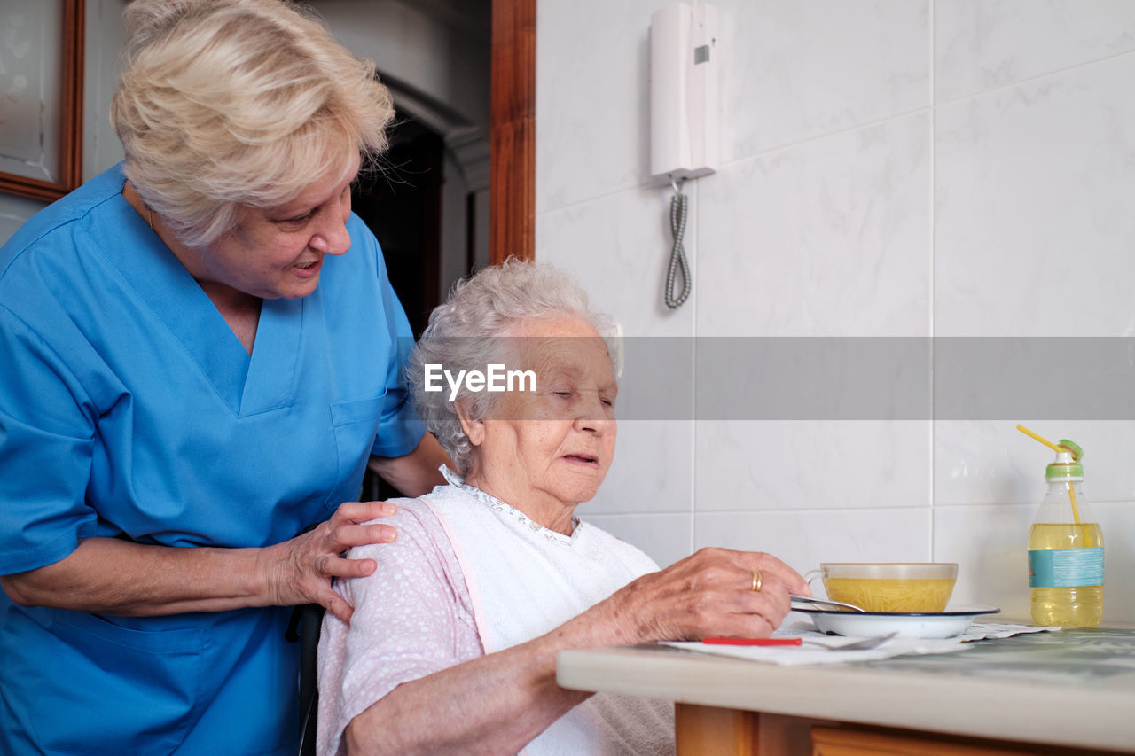 Caring nurse in uniform helping aged female patient sitting at table with bowl of soup in light kitchen at home