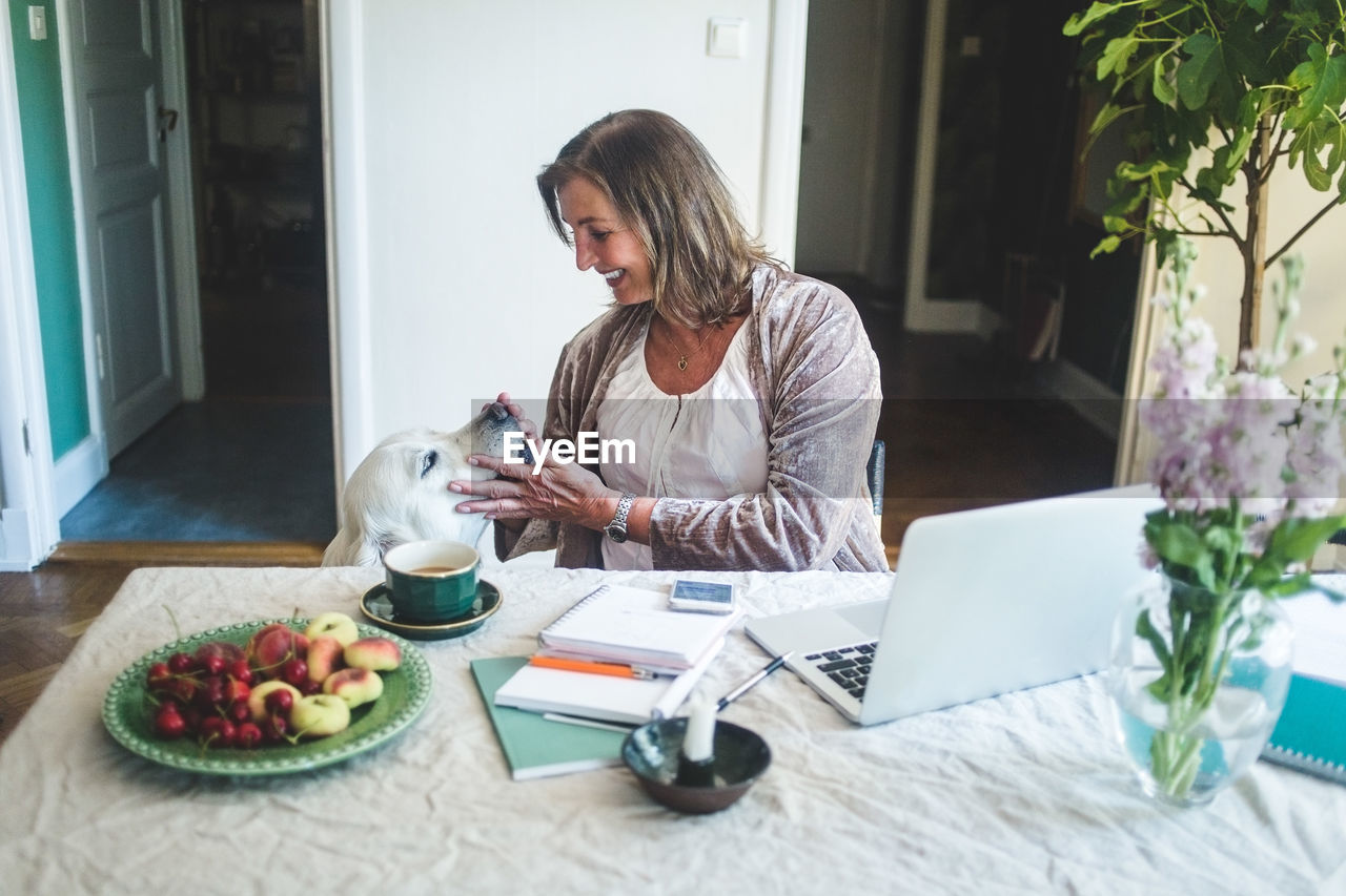 Smiling senior woman touching dog while working at table from home