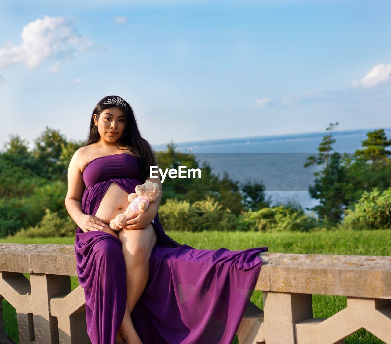 Portrait of pregnant woman with toy sitting on railing against sea