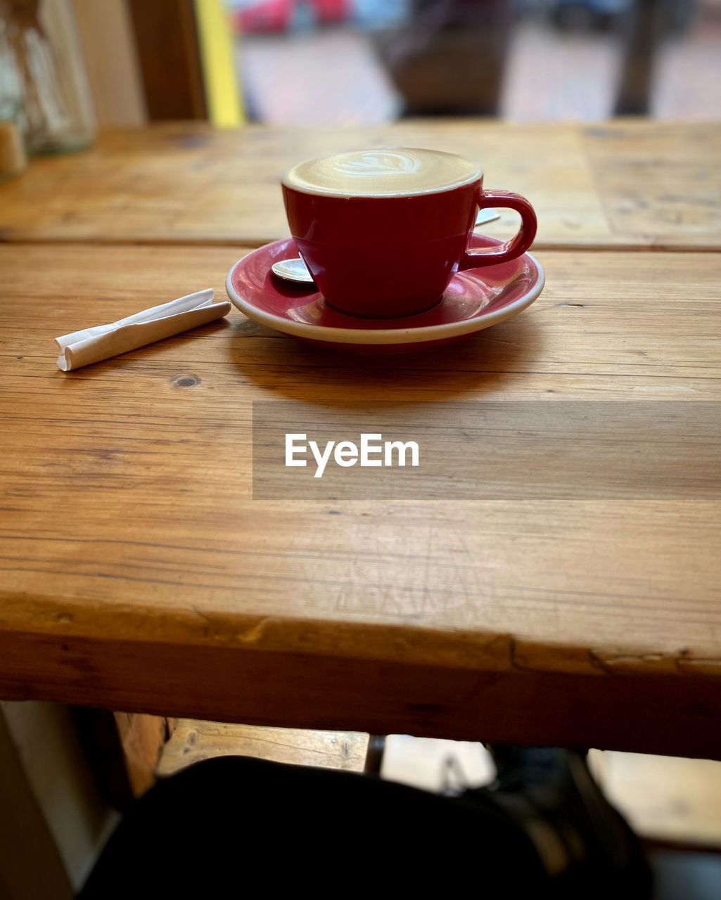 mug, cup, food and drink, table, drink, coffee cup, wood, coffee, tableware, cafe, hot drink, refreshment, indoors, crockery, saucer, spoon, kitchen utensil, focus on foreground, tea, eating utensil, furniture, seat, tea cup, restaurant, no people