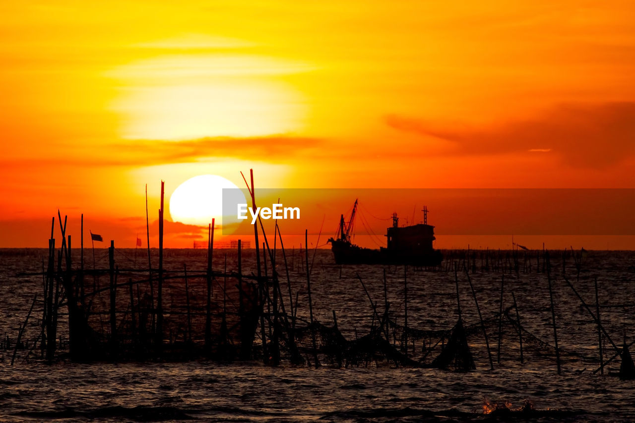 Silhouette wooden posts and ship in sea against orange sky