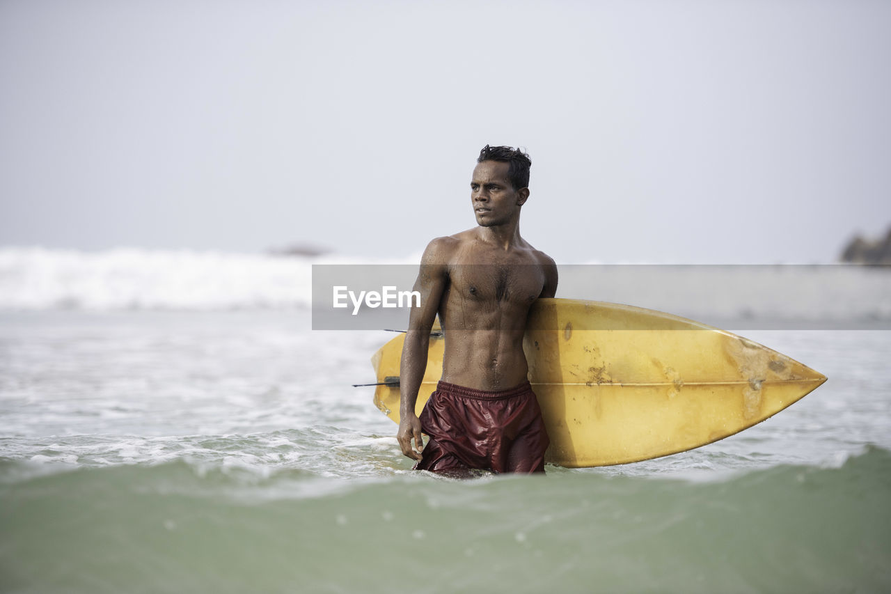 Shirtless man with surfboard standing in sea against clear sky