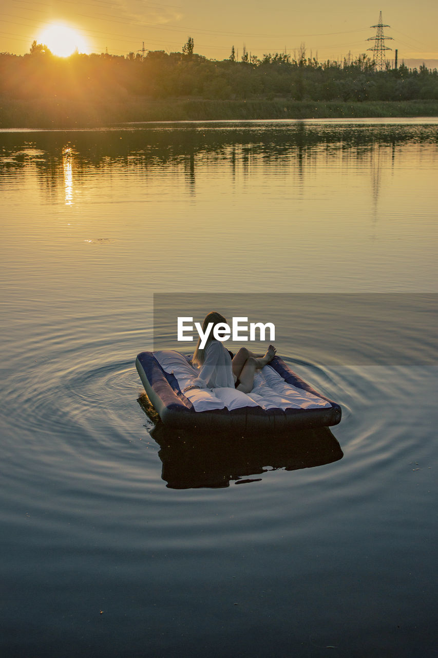 Rear view of young woman sits on a mattress on the lake during sunset