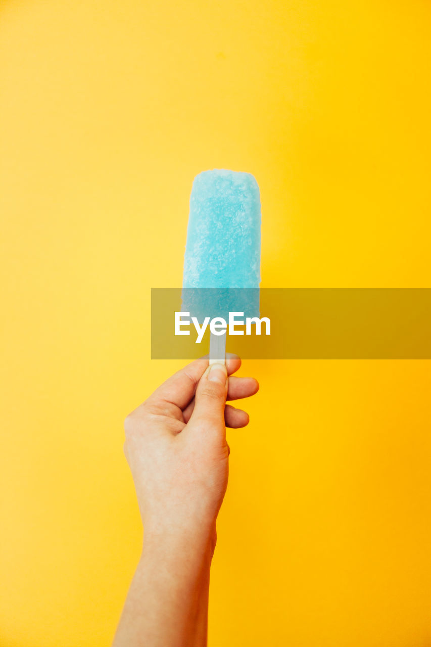 Close-up of hand holding popsicle against yellow background