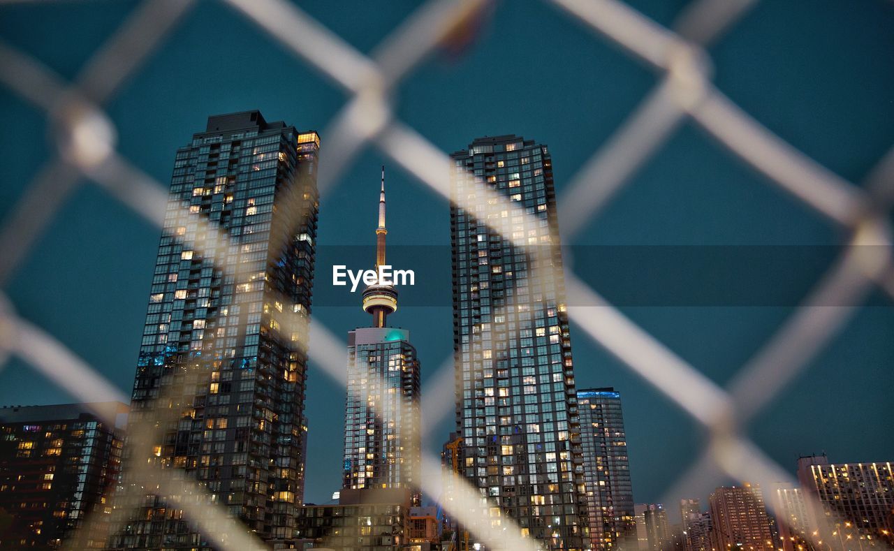 Low angle view of illuminated buildings seen through fence against sky