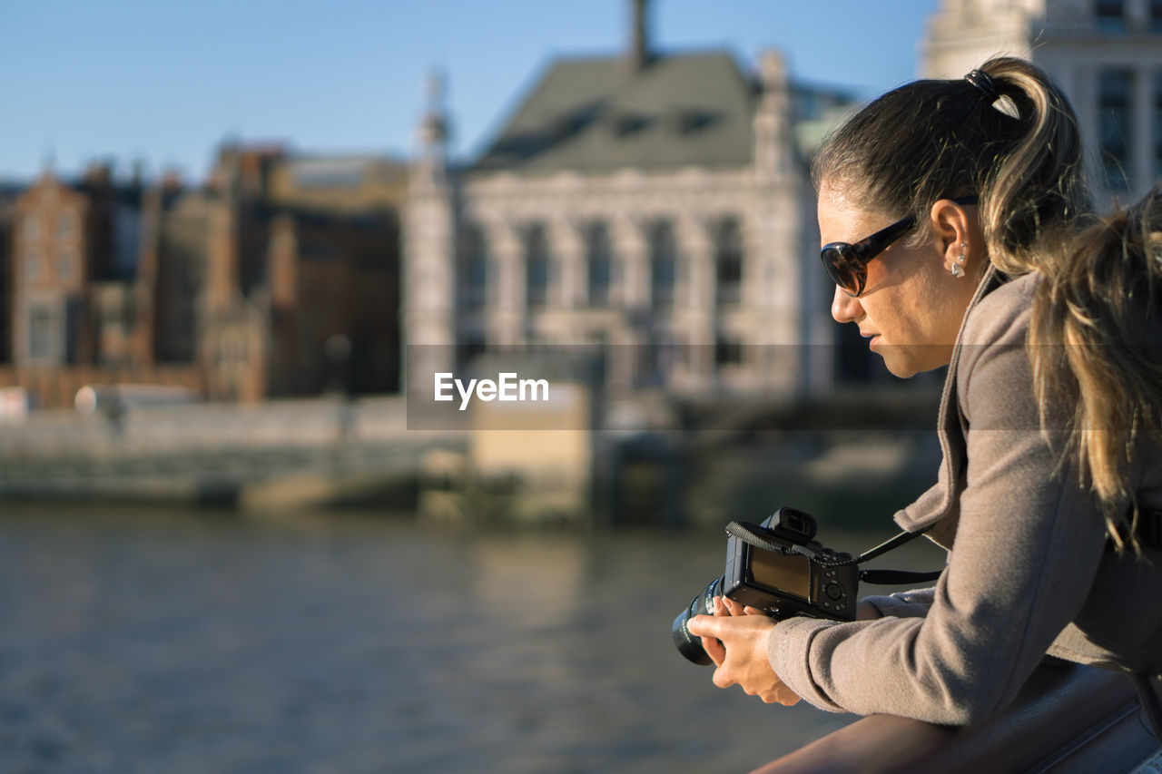 Side view of thoughtful woman with camera leaning on railing against river