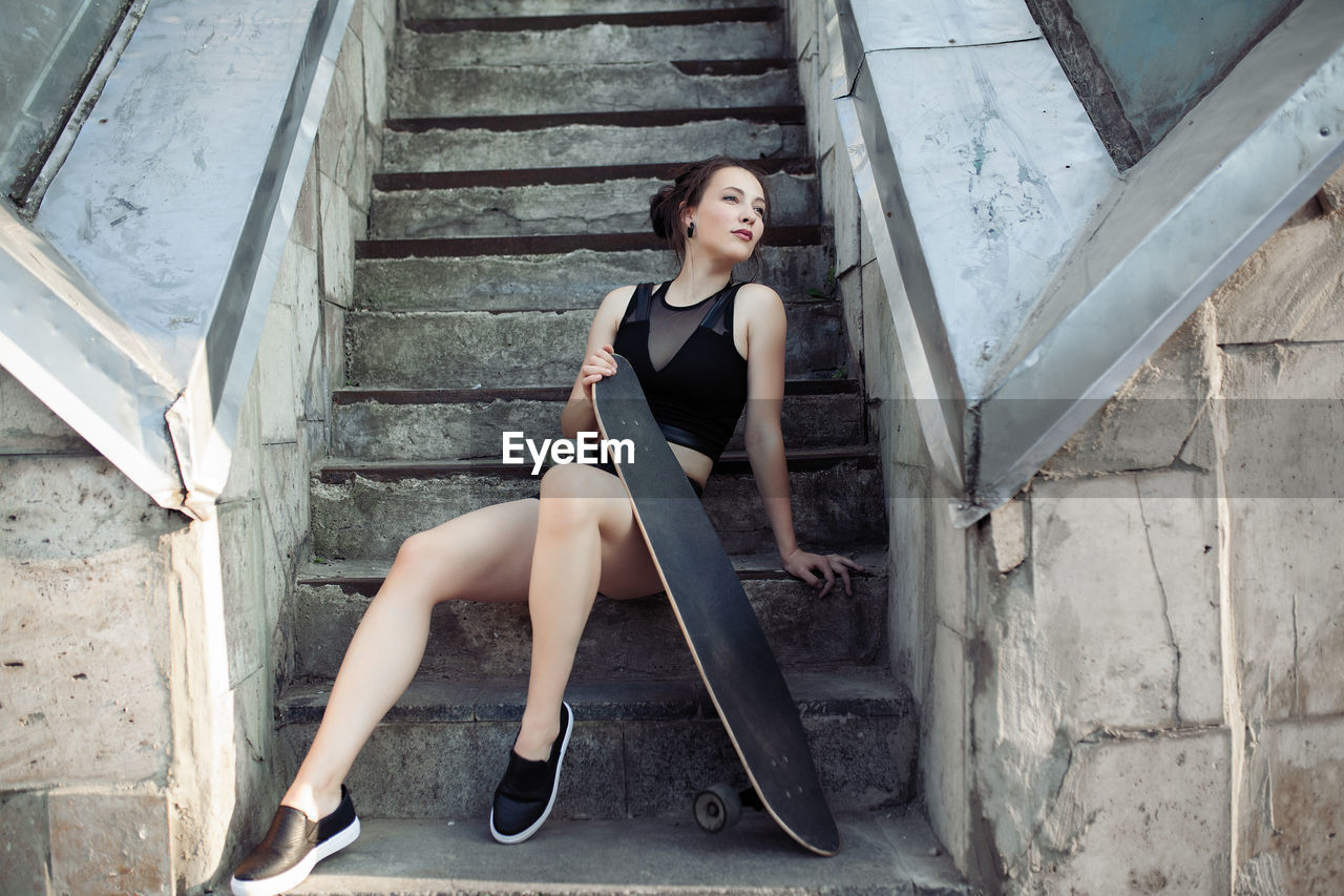 Full length of beautiful woman holding skateboard while sitting on staircase