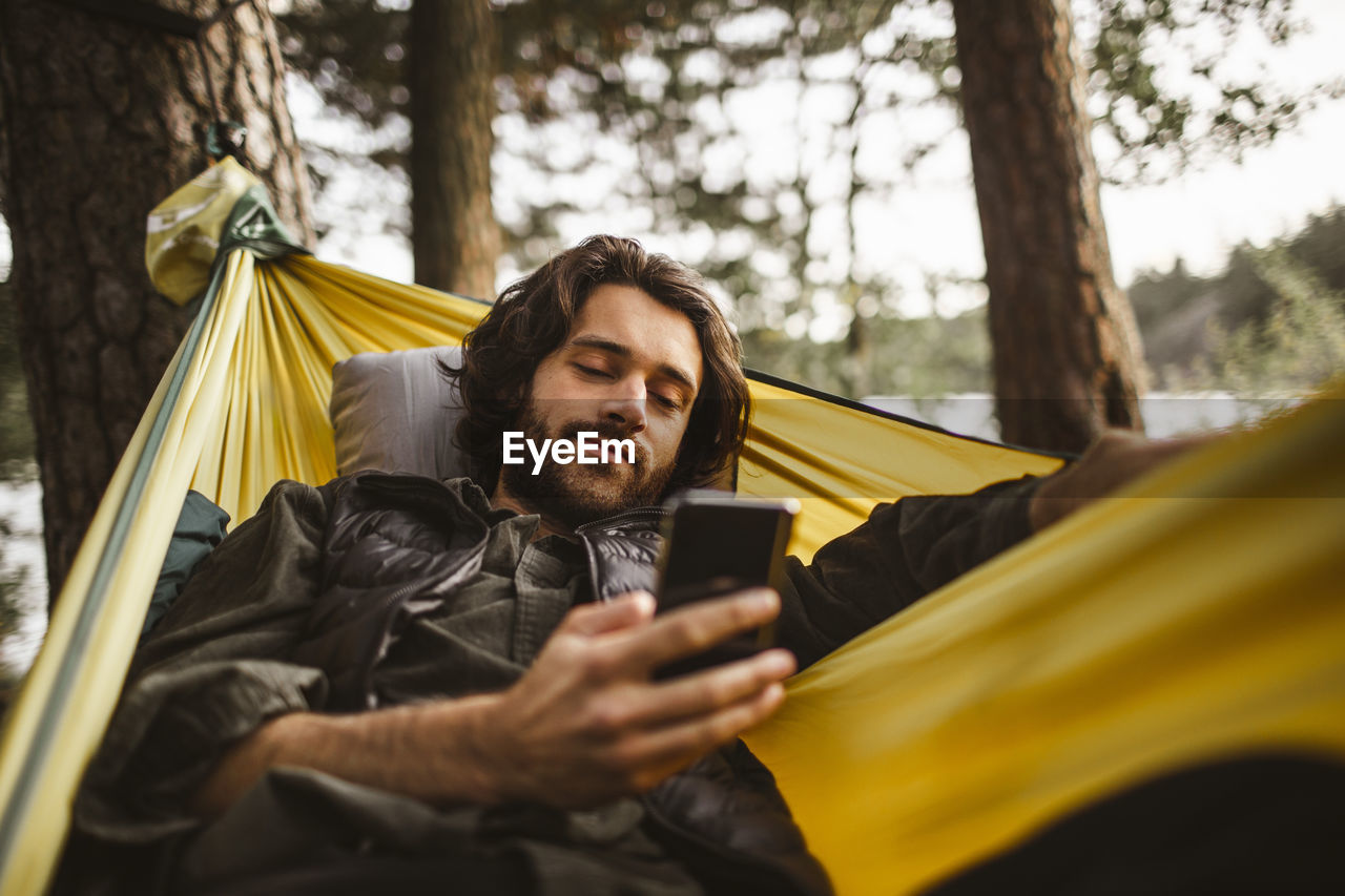 Young man using smart phone while lying over hammock in forest