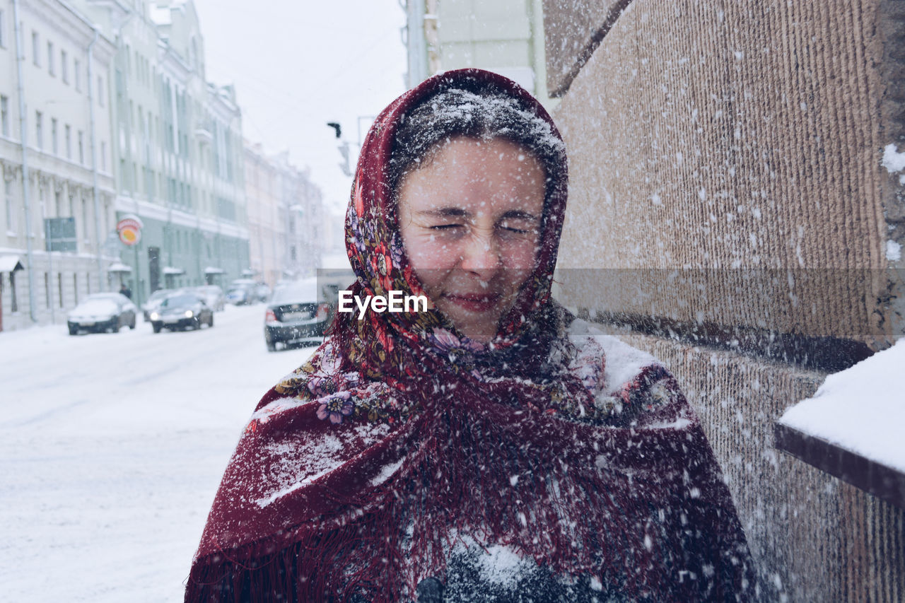 Woman with eyes closed standing by built structure in city during winter