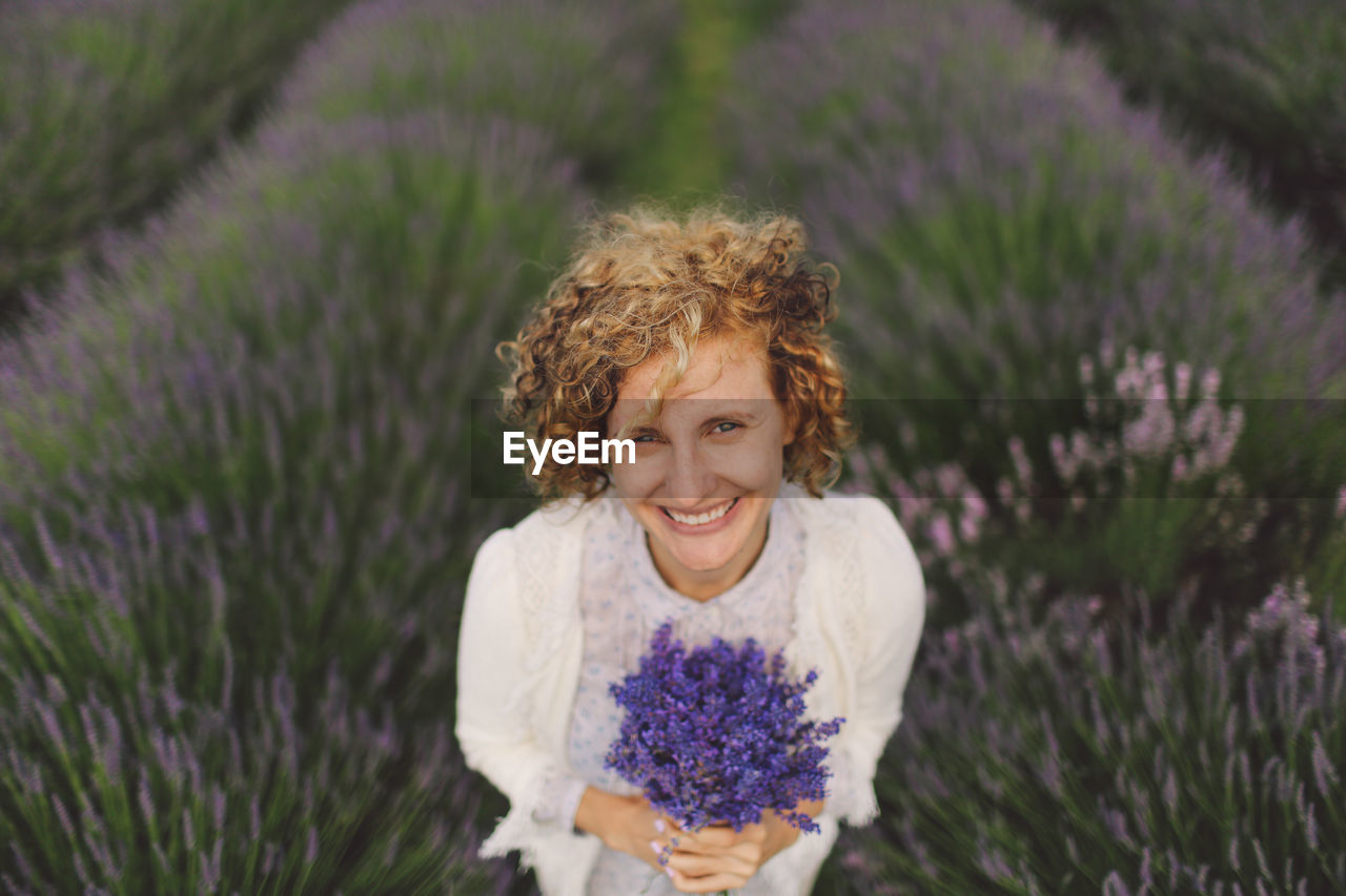 Portrait of cheerful woman holding lavender flowers on field