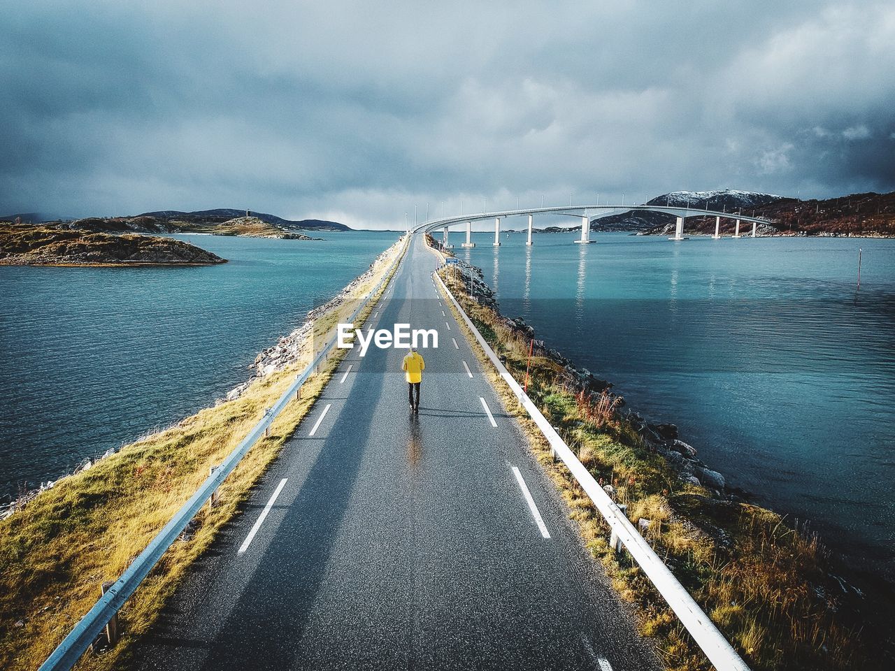 Rear view of man walking on road over sea against cloudy sky