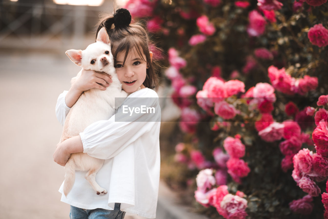 Smiling kid girl 3-4 year old holding pet dog over flower background closeup. looking at camera.