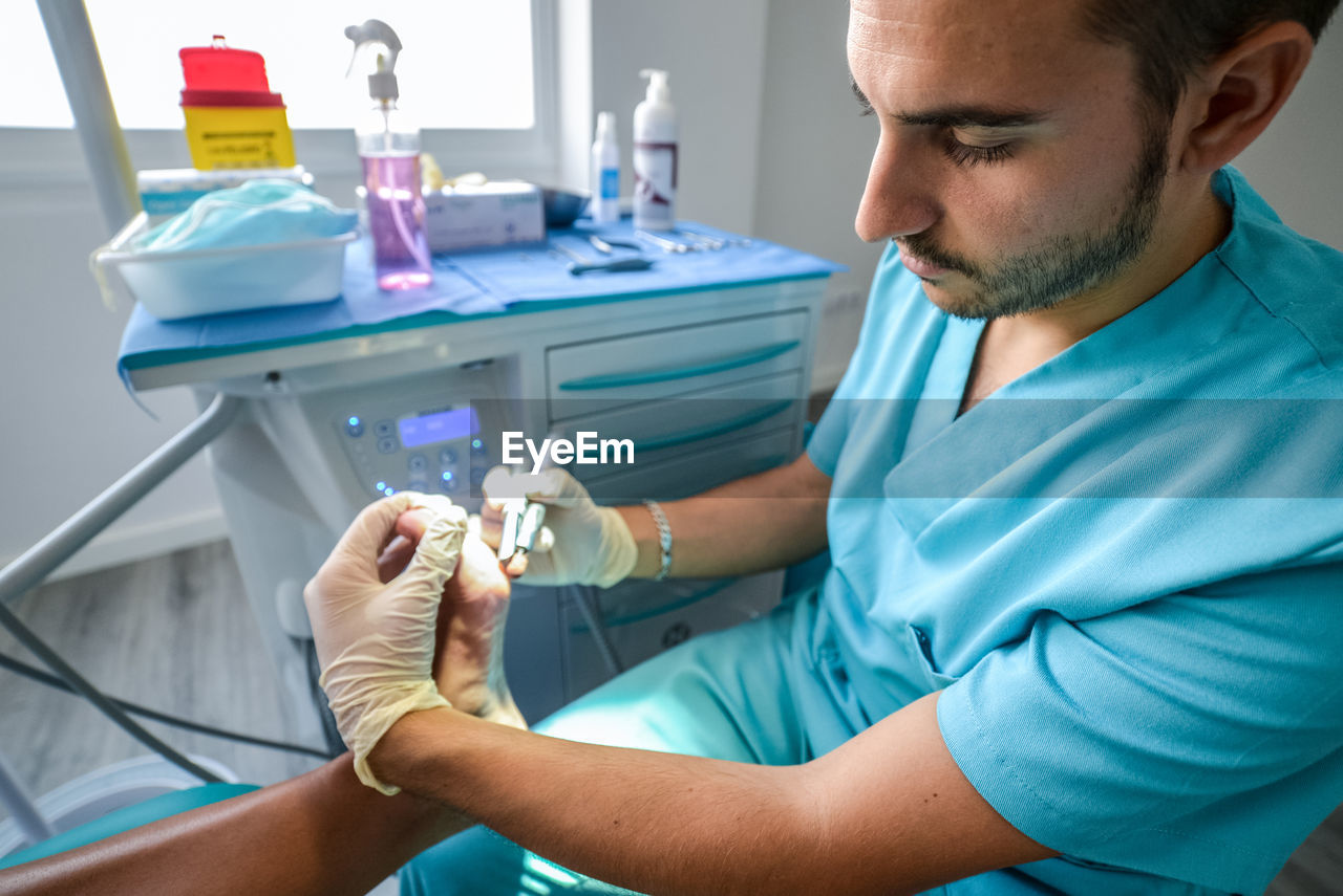Side view of qualified medical specialist in blue uniform and protective gloves using professional equipment while performing procedure with foot of patient