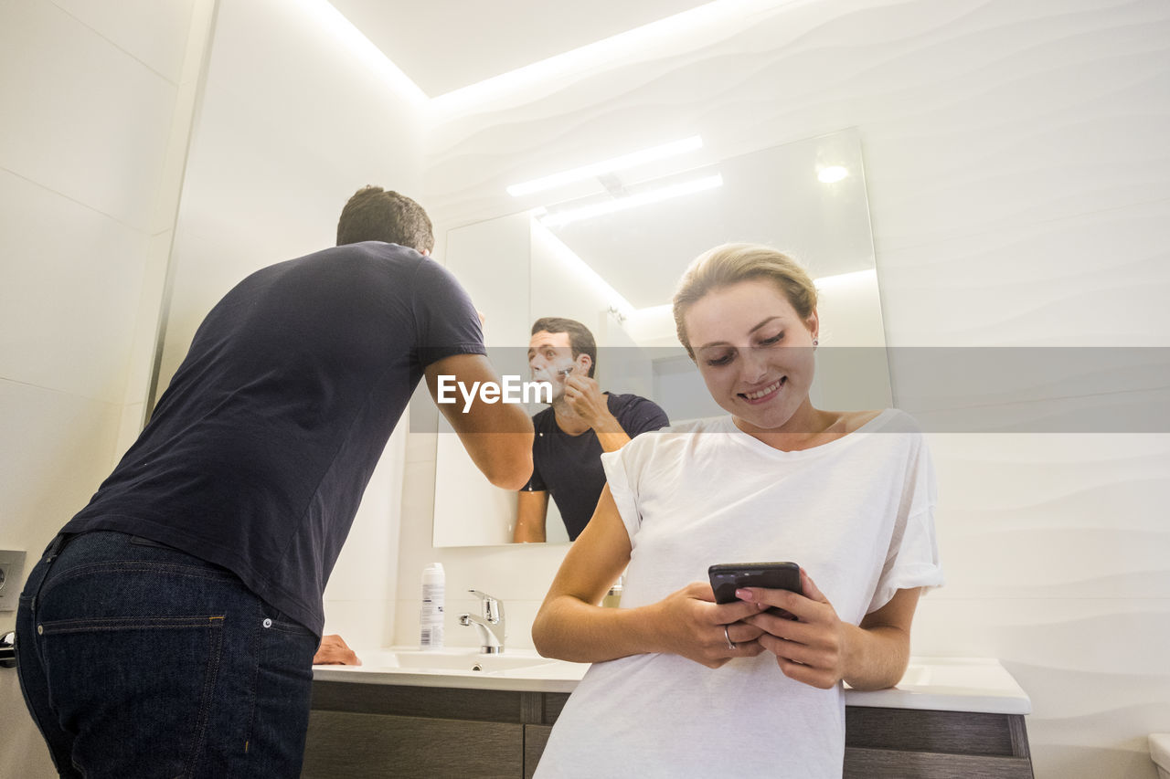 Woman using mobile phone while man shaving beard at home