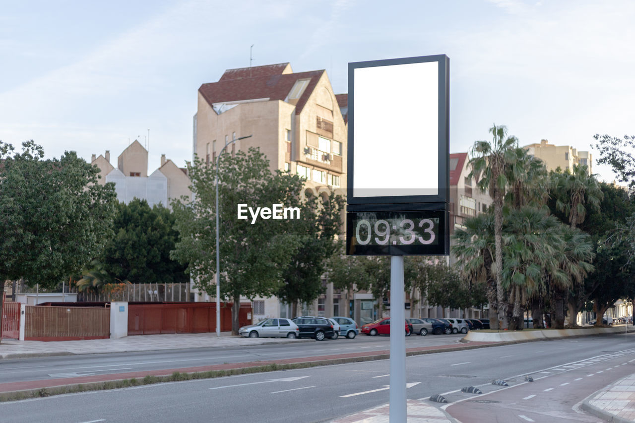 Urban digital clock post with a blank advertising panel against a cityscape, ideal for promotions. 