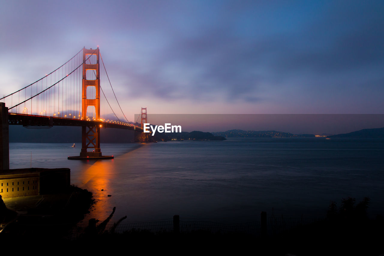 Suspension bridge over sea against cloudy sky during sunset