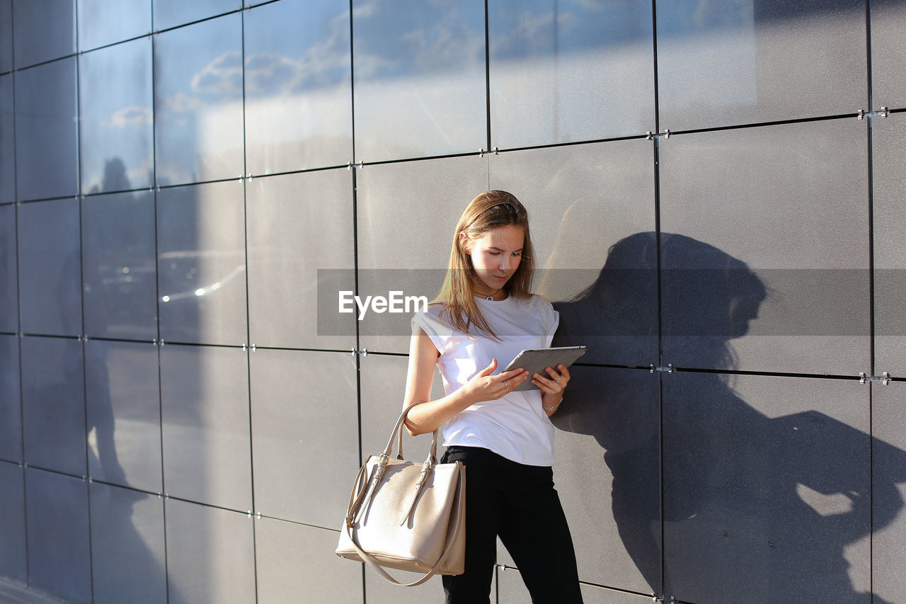 Young woman using digital tablet standing by wall