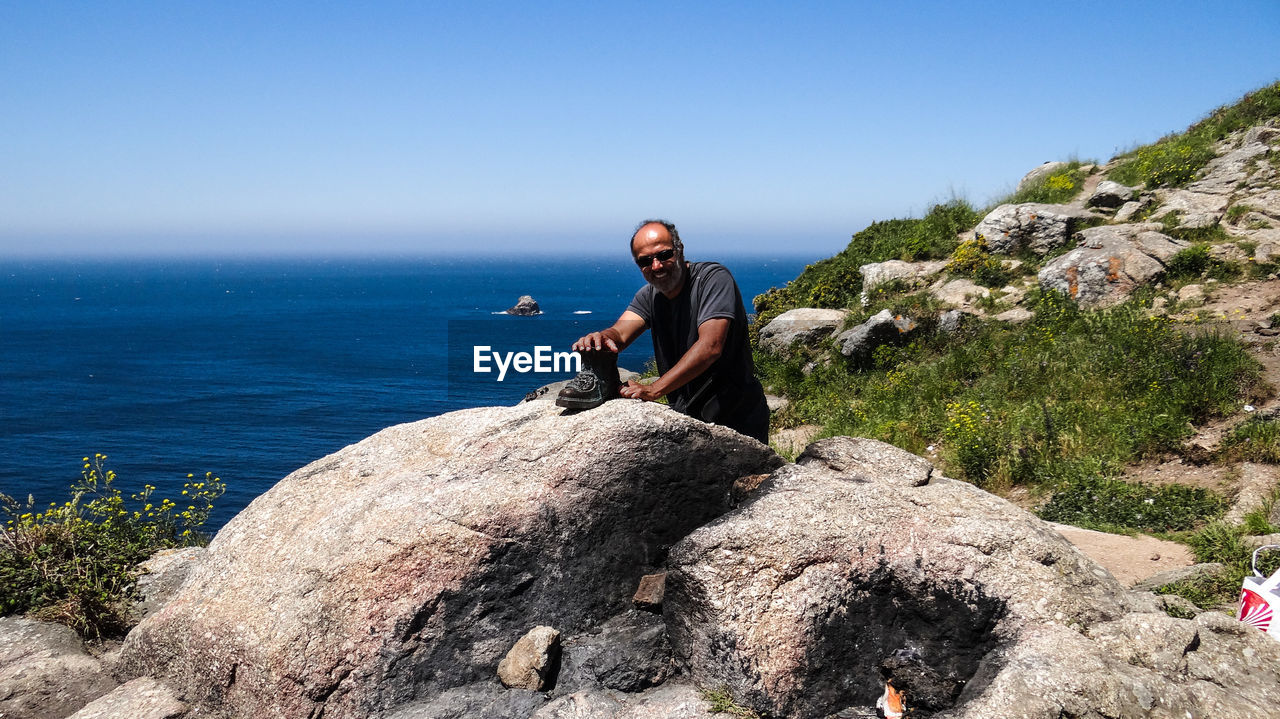 Smiling man holding boot on rock formation by blue sea against clear sky
