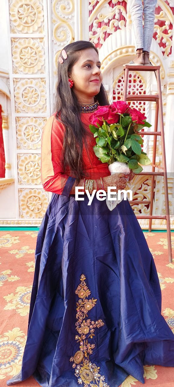 one person, women, adult, full length, clothing, smiling, young adult, flower, dress, portrait, happiness, emotion, lifestyles, costume, holding, plant, flowering plant, indoors, looking at camera, celebration, traditional clothing, person, female, sitting, fashion, nature, bride, long hair, hairstyle, gown, front view, red, brown hair, architecture, looking