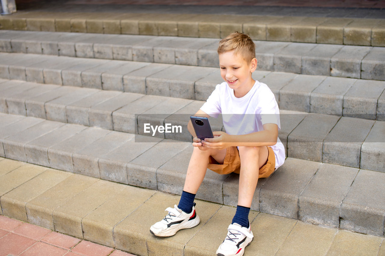 The boy is sitting on the steps near the school. play on your phone