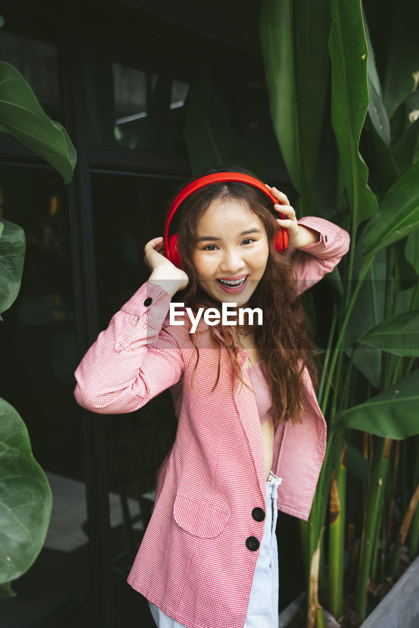 Portrait of young woman wearing headphones standing against tree