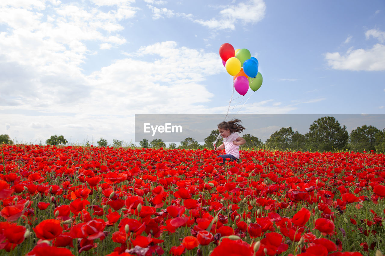 Girl holding colorful balloons jumping amidst red poppy flowers against sky