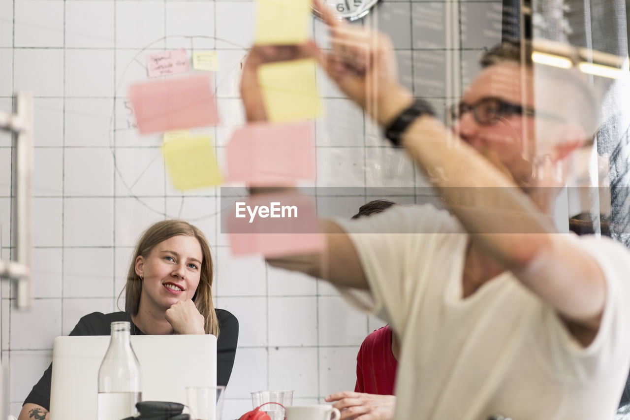 Young businessman writing ideas on adhesive notes with colleagues in background at creative office