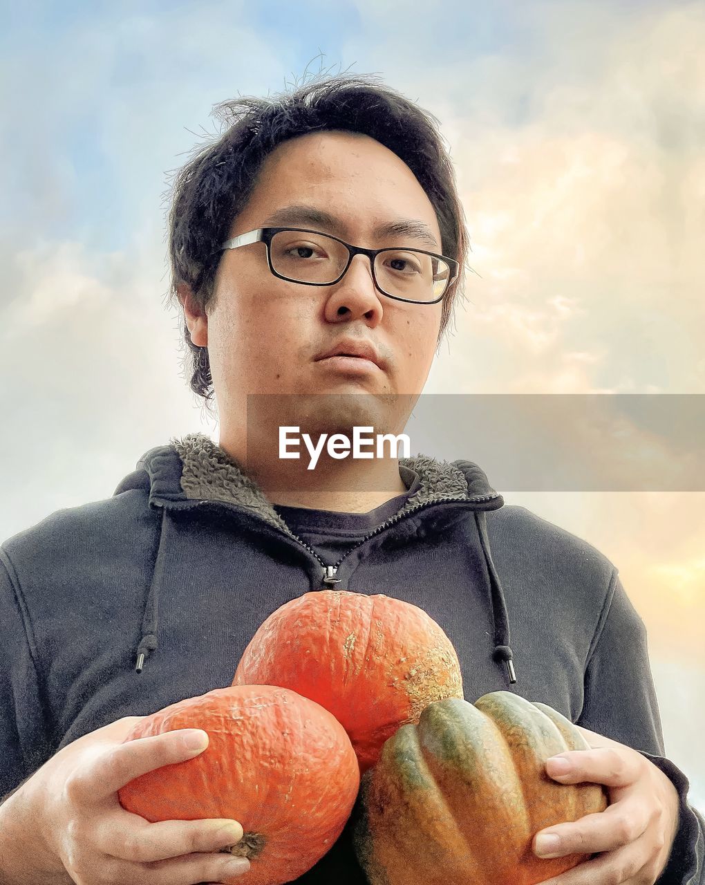 Portrait of young man holding three pumpkins against cloudy sunset sky.