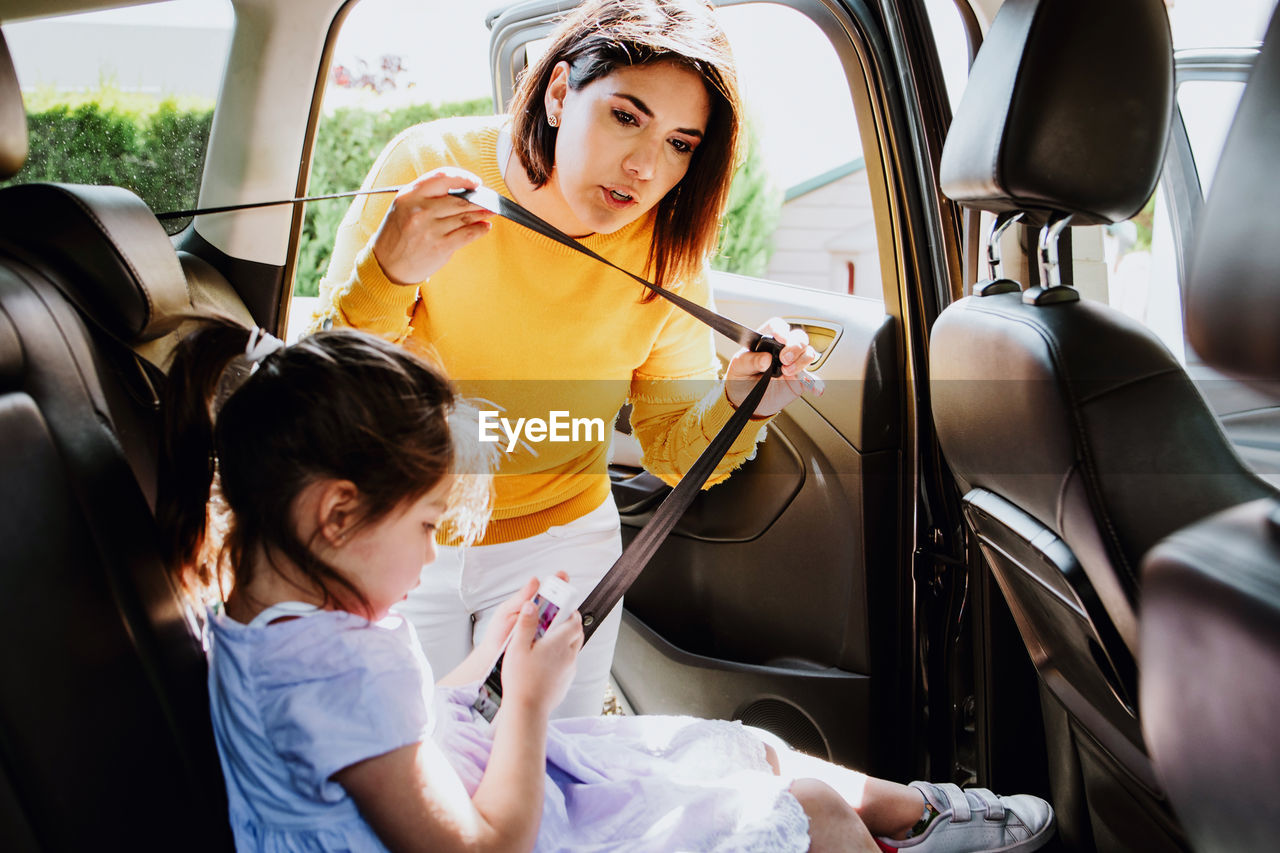 Young ethnic mother in stylish outfit fastening safety belt to little daughter using mobile phone on backseat of modern car on sunny day