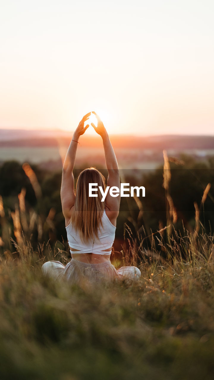 Back view on woman sitting in meditation yoga pose and catching sun by hands at sunset outdoors	
