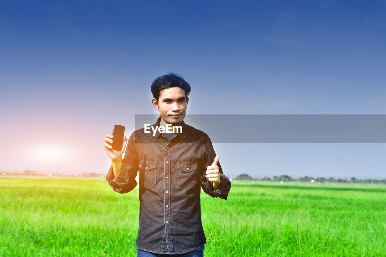 Portrait of man showing smart phone while standing on field against sky