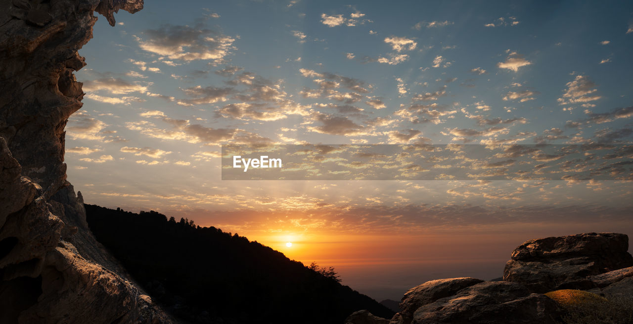 Silhouette view from top of mountains at sunrise with cloudy sky and cliff in foreground, 