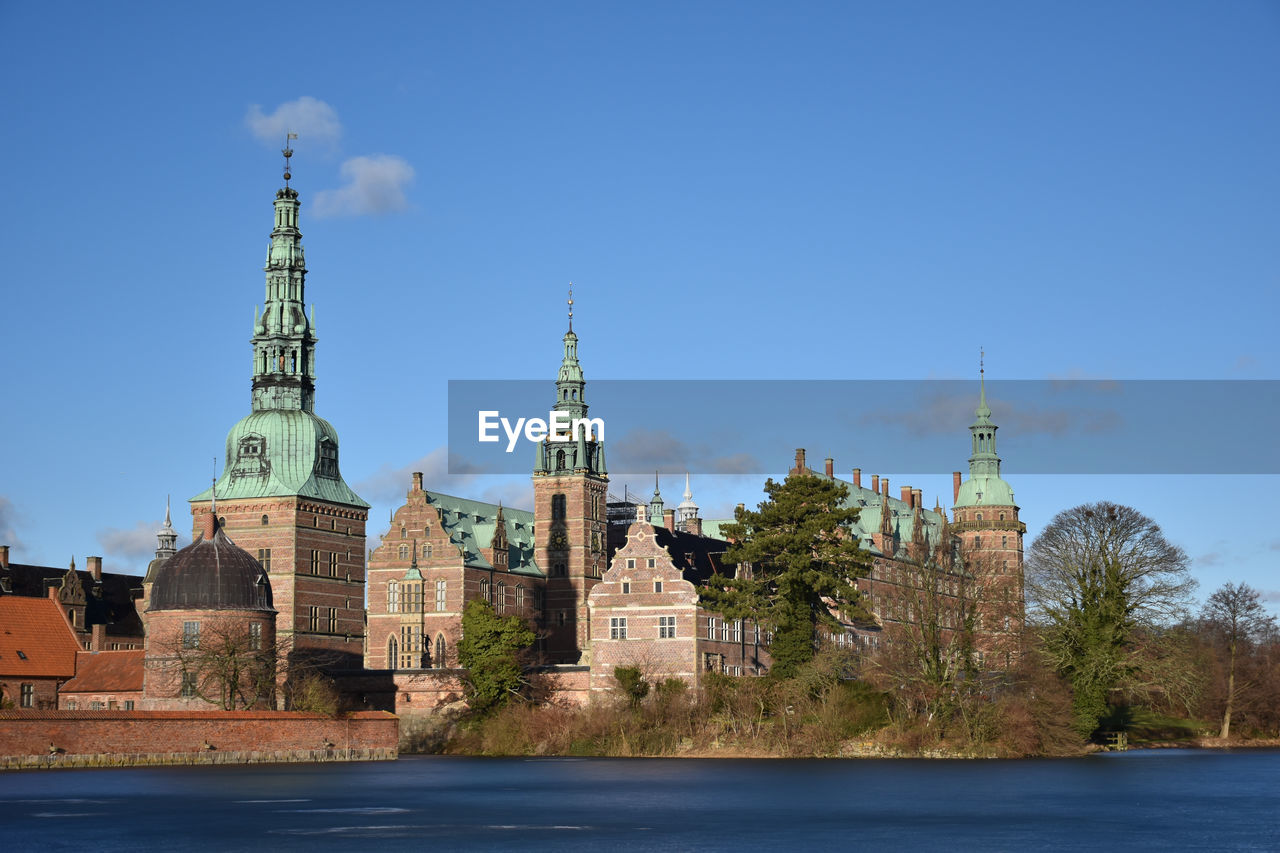 Frederiksborg castle by river against blue sky during sunny day