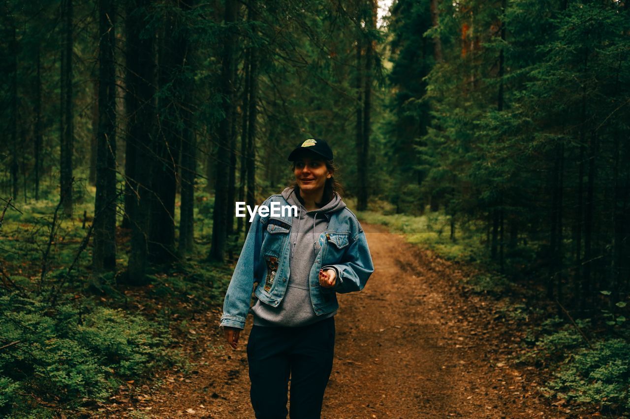 Portrait of smiling woman wearing cap walking on dirt road amidst forest