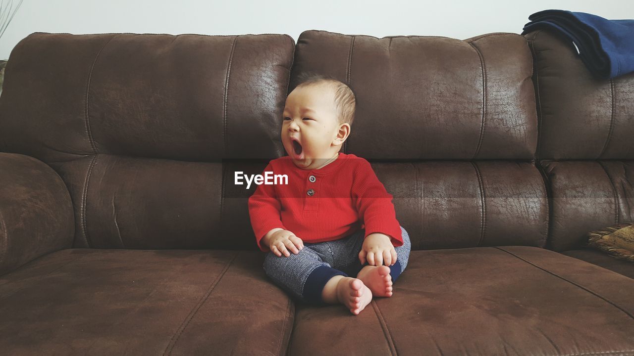 Cute baby boy yawning while sitting on sofa at home