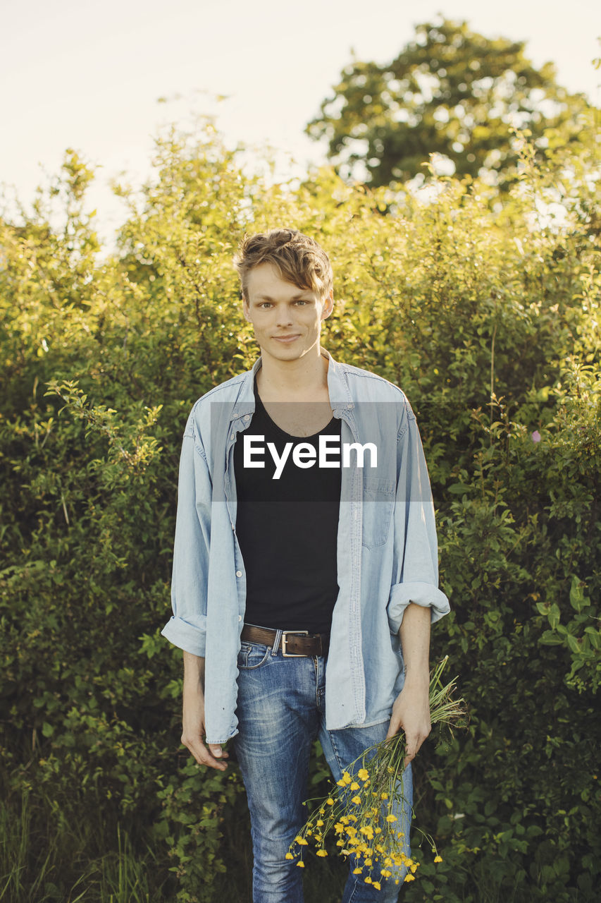 Portrait of young man holding flowers standing against plants