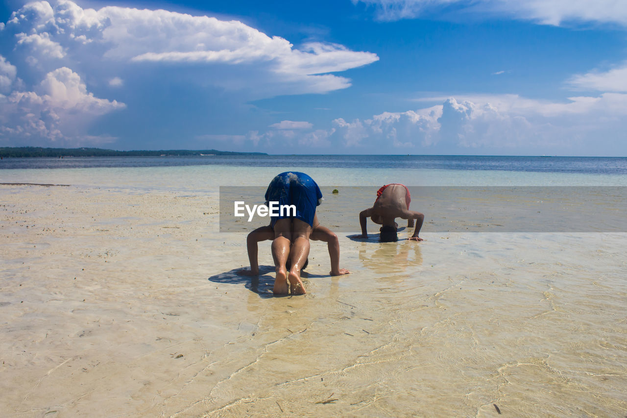 Men practicing headstand at beach against sky