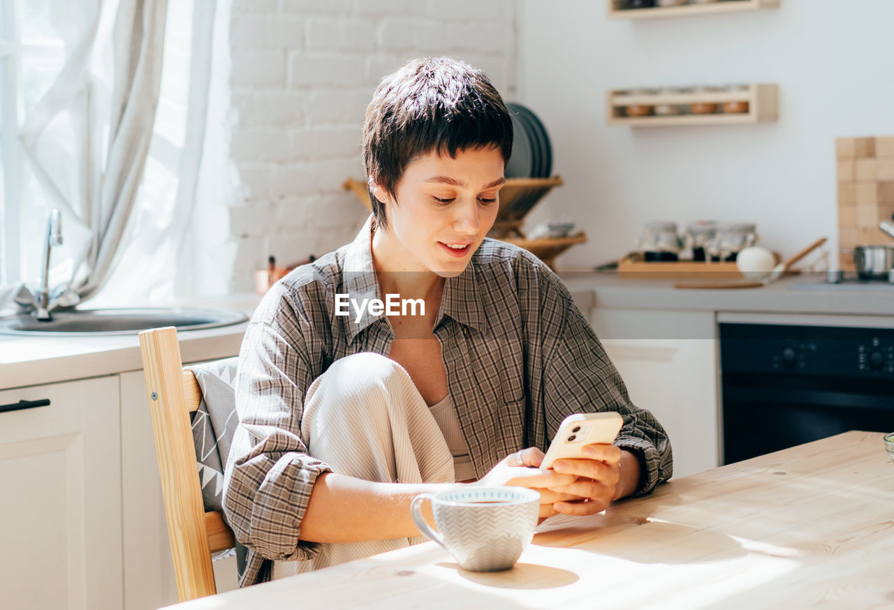 Young woman surfs the phone while sitting in the kitchen with coffee.