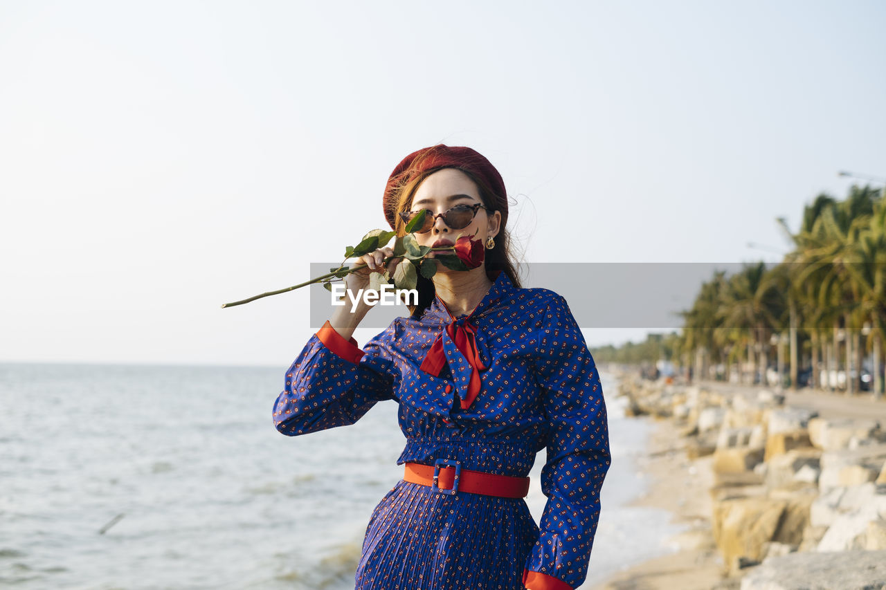 Portrait of beautiful young woman holding rose standing on beach against sky