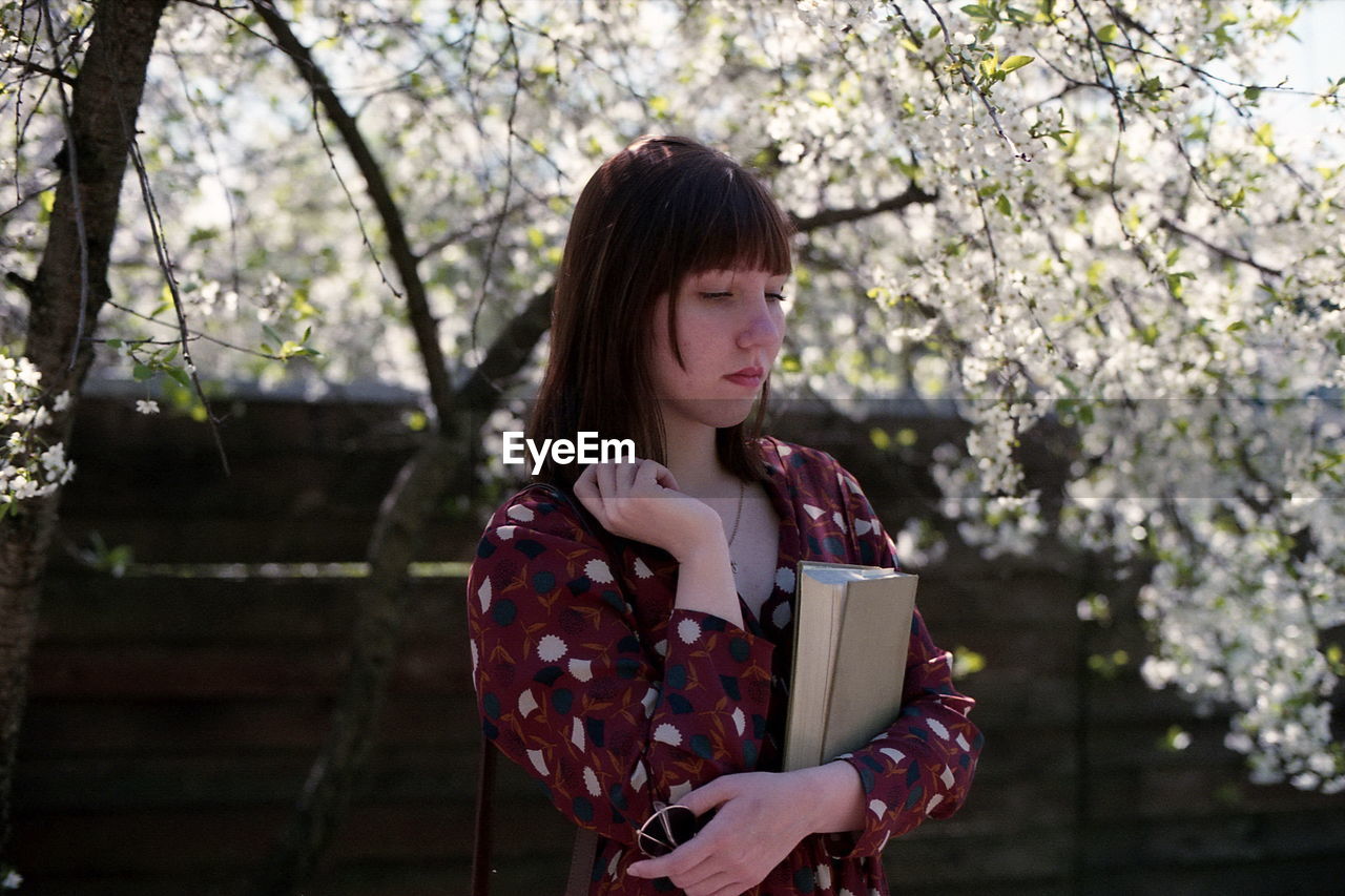 Woman holding book while standing by tree