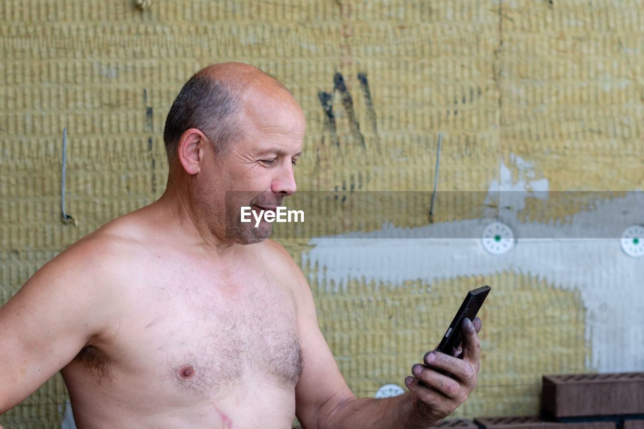 Shirtless mature man using smart phone while standing against wall