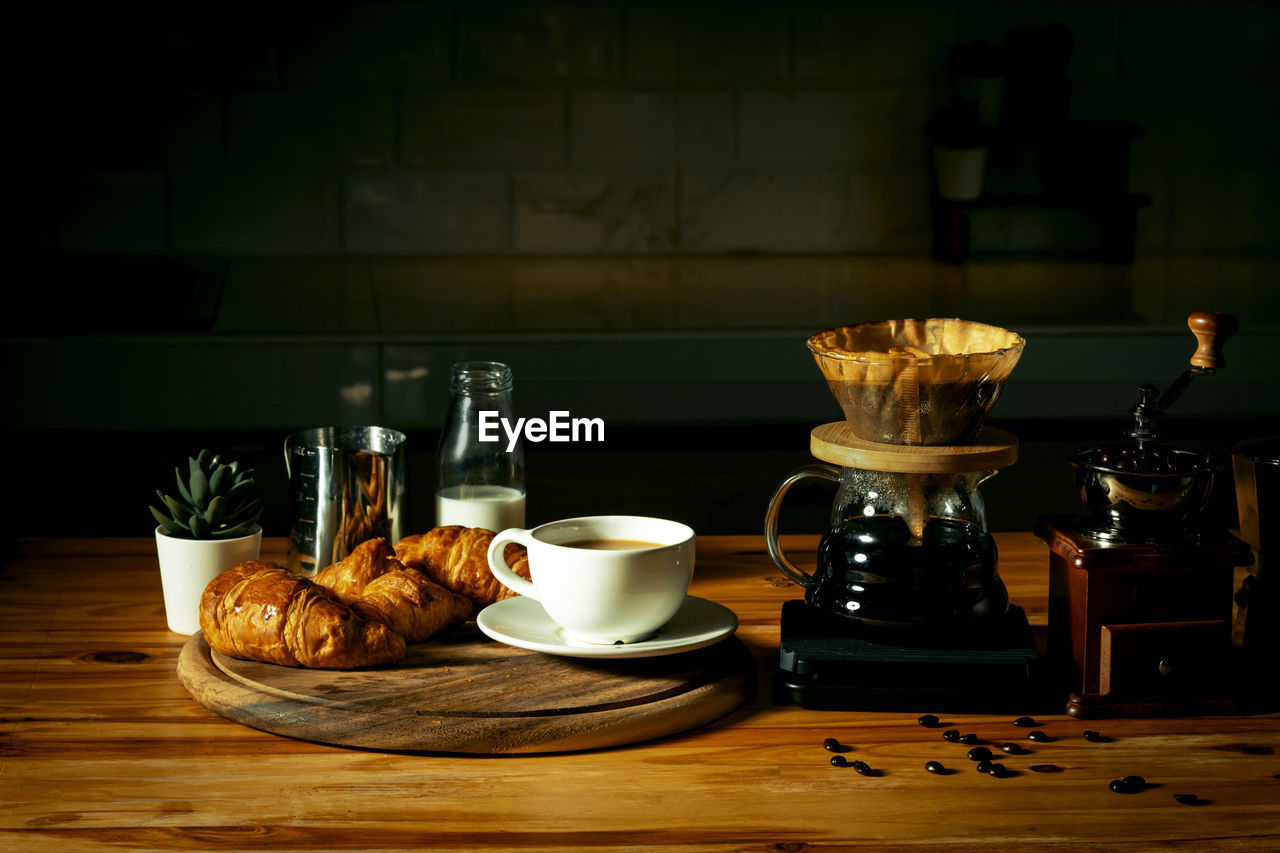 Set of drip coffee maker and fresh croissant for breakfast on dark rustic wooden table in a cafe.